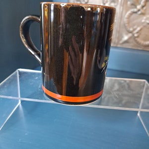 May include: Black ceramic mug with an orange stripe around the top. The mug has a design of a bird with yellow and green accents.