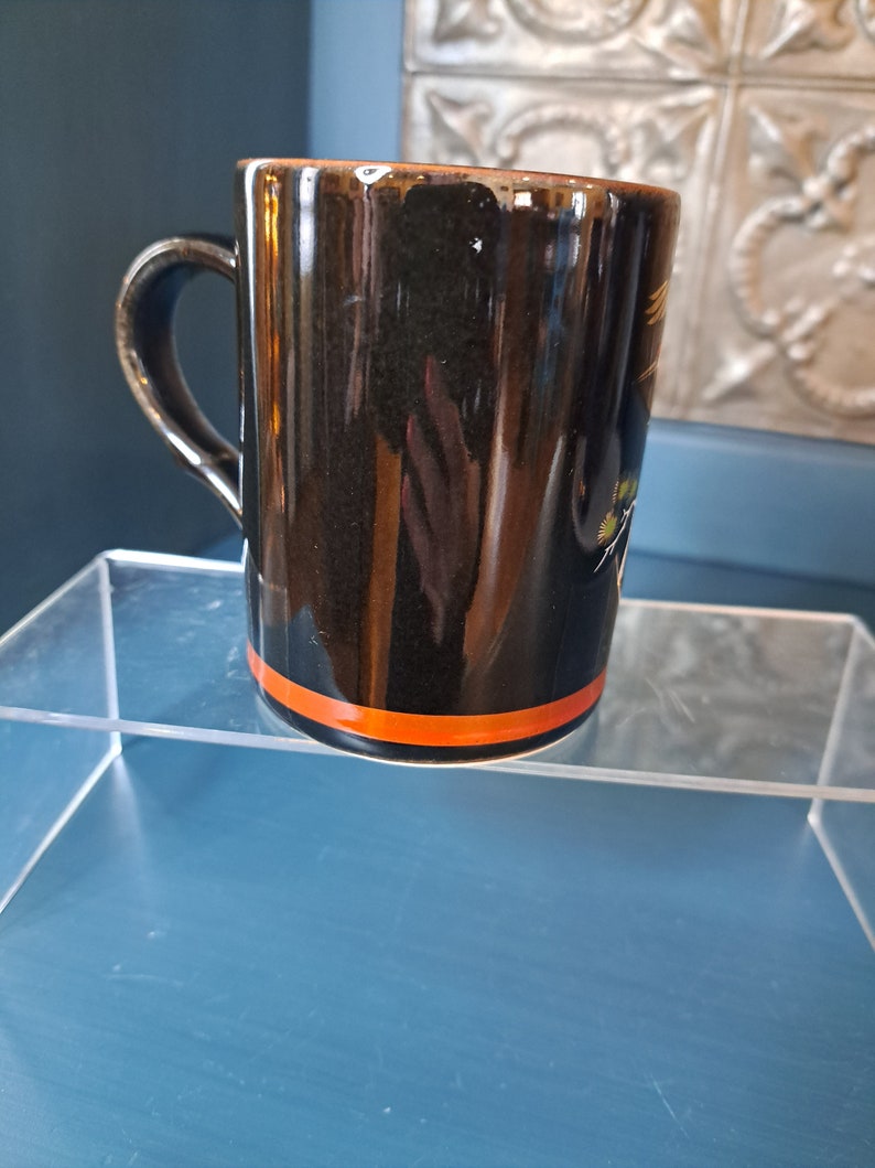 May include: Black ceramic mug with an orange stripe around the top. The mug has a design of a bird with yellow and green accents.