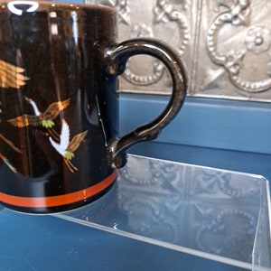 May include: Black ceramic mug with a red stripe and a gold and white crane design. The mug has a black handle.