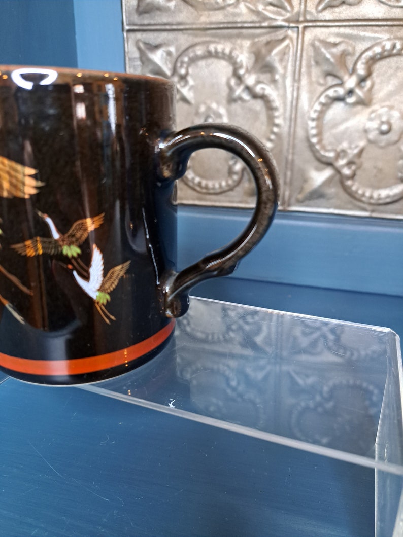 May include: Black ceramic mug with a red stripe and a gold and white crane design. The mug has a black handle.