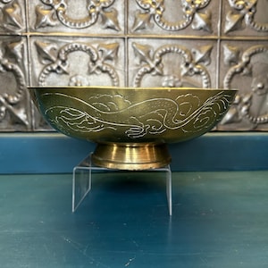 May include: A brass bowl with a dragon design etched around the rim. The bowl sits on a short, round base and is displayed on a clear acrylic stand. The background features a decorative, metallic wall.