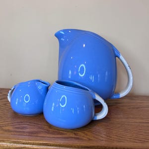 May include: A blue ceramic pitcher and two matching mugs with white and blue handles. The pitcher has a curved spout and a large handle. The mugs are round with a handle. The set is displayed on a wooden surface.