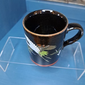 May include: Black ceramic mug with a red rim and a design of white cranes flying with green and orange accents.