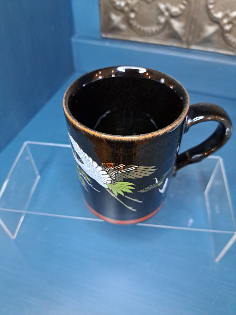 May include: Black ceramic mug with a red rim and a design of white cranes flying with green and orange accents.