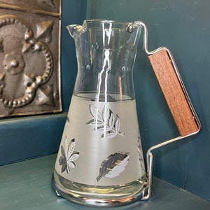 May include: A clear glass pitcher with a silver frosted design of leaves. The pitcher has a chrome base and a wooden handle.