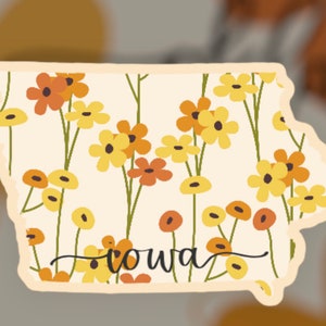 Puede incluir: Una pegatina del estado de Iowa con un patrón floral en tonos amarillos, naranjas y verdes. La palabra "Iowa" está escrita en cursiva en la parte inferior de la pegatina.