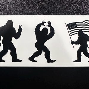 May include: A black and white sticker featuring three Bigfoot silhouettes. One gives a peace sign, another forms a heart with its hands, and the third carries an American flag. The sticker is on a white background.