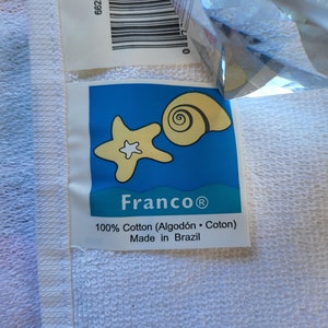 May include: A white towel with a blue label featuring a starfish and seashell. The label reads "Franco&reg; 100% Cotton (Algod&oacute;n &middot; Coton) Made in Brazil".