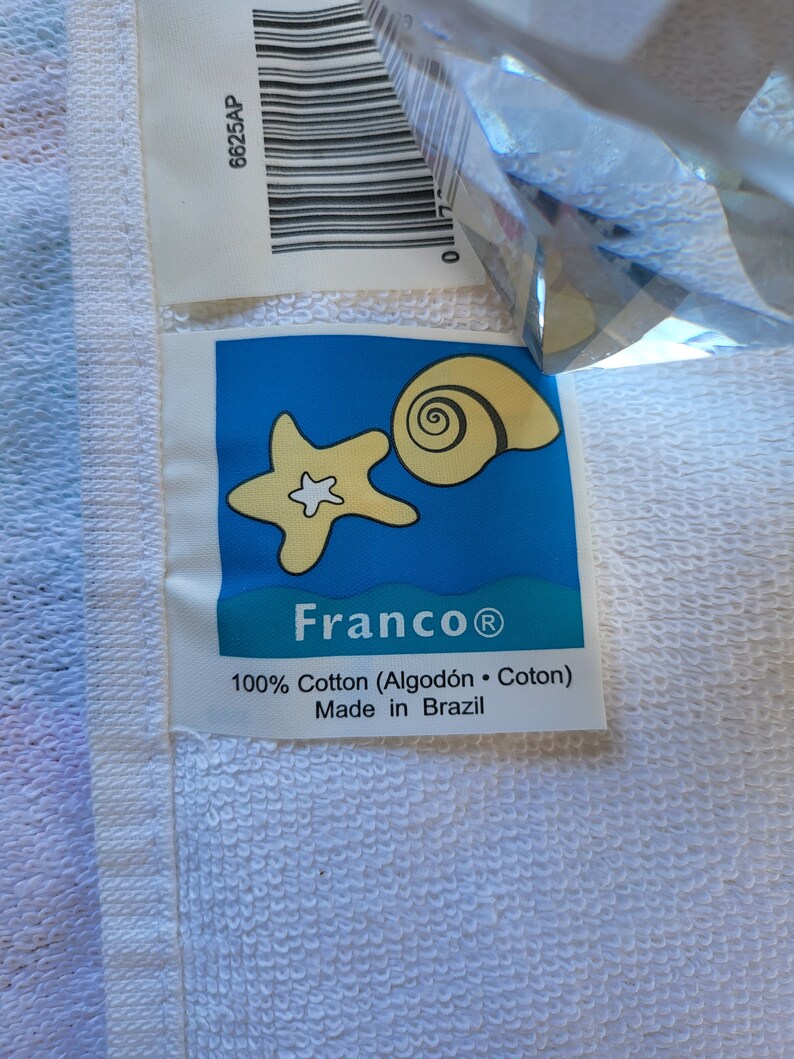 May include: A white towel with a blue label featuring a starfish and seashell. The label reads "Franco&reg; 100% Cotton (Algod&oacute;n &middot; Coton) Made in Brazil".