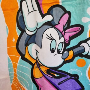 May include: A colorful beach towel featuring a cartoon image of Minnie Mouse wearing a pink dress and a purple bag. The background is a mix of orange, blue, and white swirls.