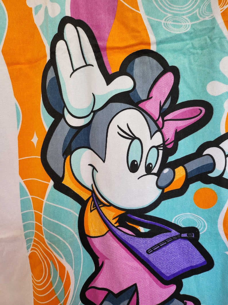 May include: A colorful beach towel featuring a cartoon image of Minnie Mouse wearing a pink dress and a purple bag. The background is a mix of orange, blue, and white swirls.