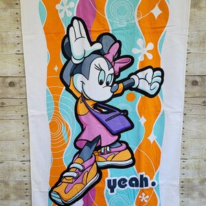 May include: A white beach towel with a colorful design featuring Minnie Mouse in a pink outfit and sneakers. The background is a mix of orange, blue, and pink swirls with the word "yeah" in black text. The towel is made of soft, absorbent material.