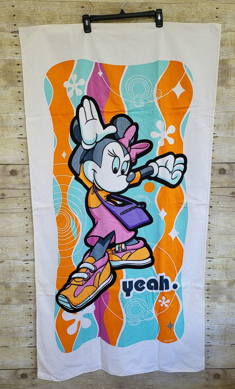 May include: A white beach towel with a colorful design featuring Minnie Mouse in a pink outfit and sneakers. The background is a mix of orange, blue, and pink swirls with the word "yeah" in black text. The towel is made of soft, absorbent material.