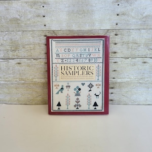 May include: A red book titled "Historic Samplers" with a white background and black and white illustrations of birds, trees, and other designs. The book cover also includes the alphabet and numbers.