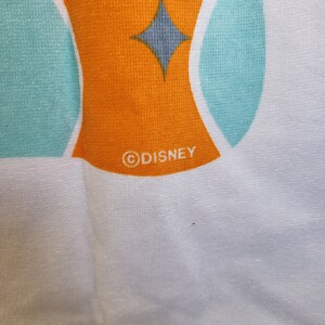 May include: White beach towel with a turquoise and orange geometric design. The towel has a gray diamond shape and the word "DISNEY" in a circle.