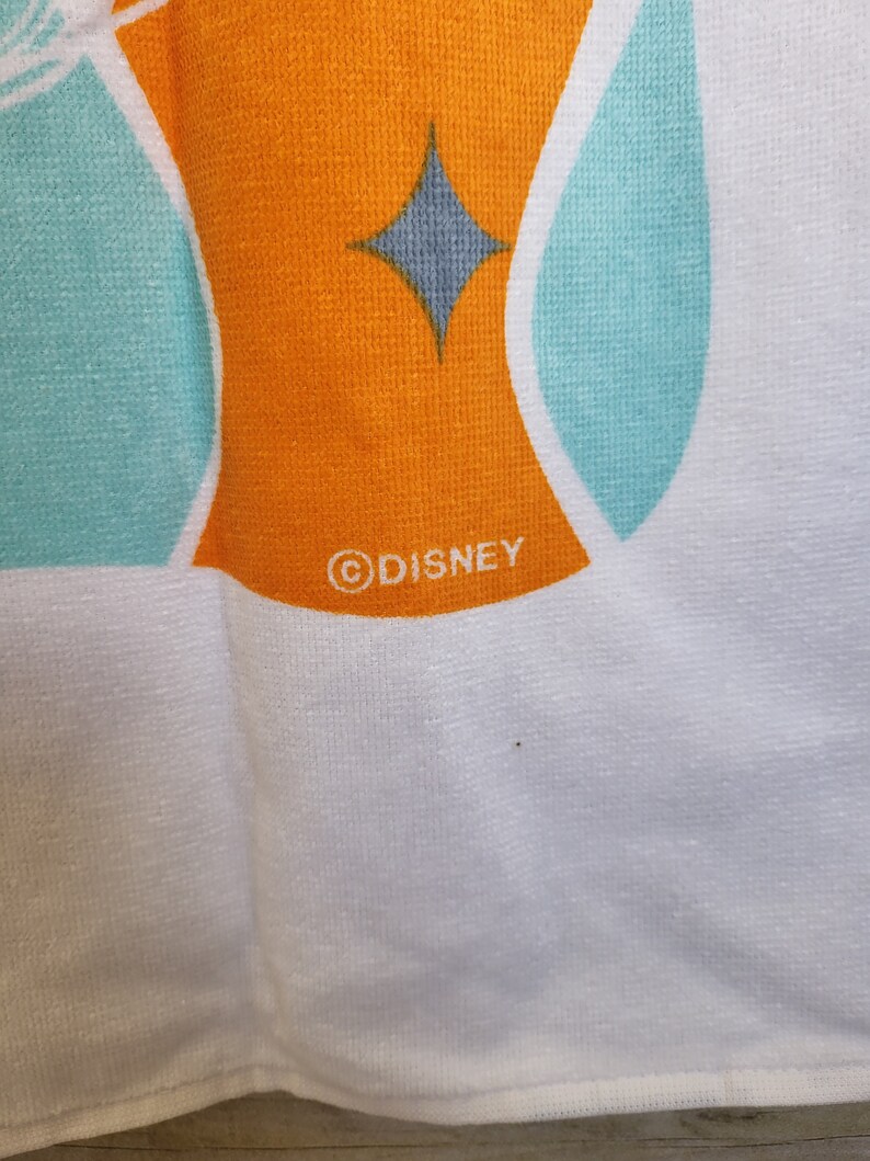 May include: White beach towel with a turquoise and orange geometric design. The towel has a gray diamond shape and the word "DISNEY" in a circle.
