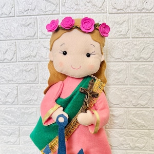 May include: A handmade felt doll of a saint with a pink dress, green robe, and a blue anchor. The doll is wearing a crown of pink flowers and has brown hair.