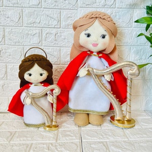 May include: Two felt dolls dressed in white robes with red capes and gold trim, holding gold harps. The dolls have brown hair and painted faces.
