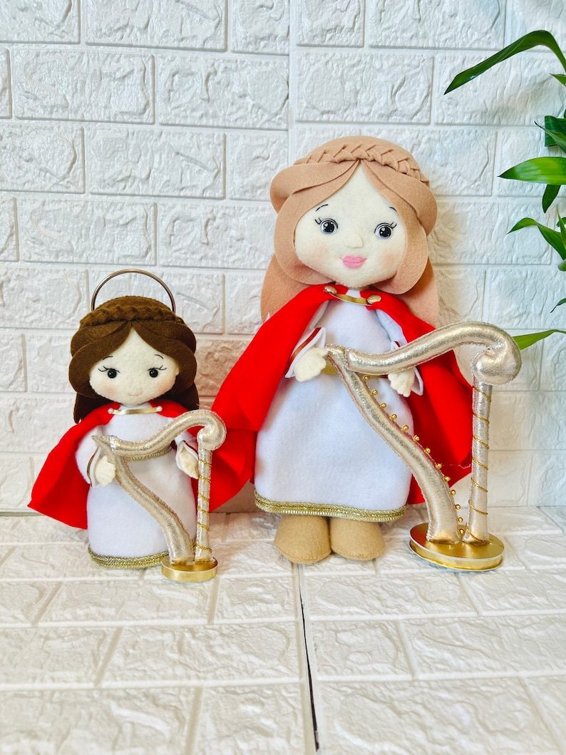 May include: Two felt dolls dressed in white robes with red capes and gold trim, holding gold harps. The dolls have brown hair and painted faces.