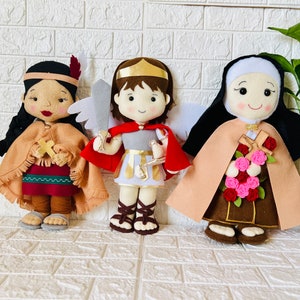 May include: Three felt dolls, each with a different design and color scheme. The doll on the left is wearing a brown and red outfit with a feather headdress. The doll in the middle is wearing a red and white outfit with a gold crown. The doll on the right is wearing a brown and beige outfit with a white head covering.