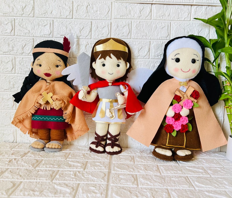 May include: Three felt dolls, each with a different design and color scheme. The doll on the left is wearing a brown and red outfit with a feather headdress. The doll in the middle is wearing a red and white outfit with a gold crown. The doll on the right is wearing a brown and beige outfit with a white head covering.