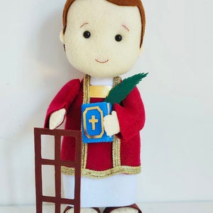May include: A felt doll of a religious figure wearing a red robe with gold trim, holding a book with a cross on it and a palm branch. The doll is standing next to a brown felt cutout of a window.