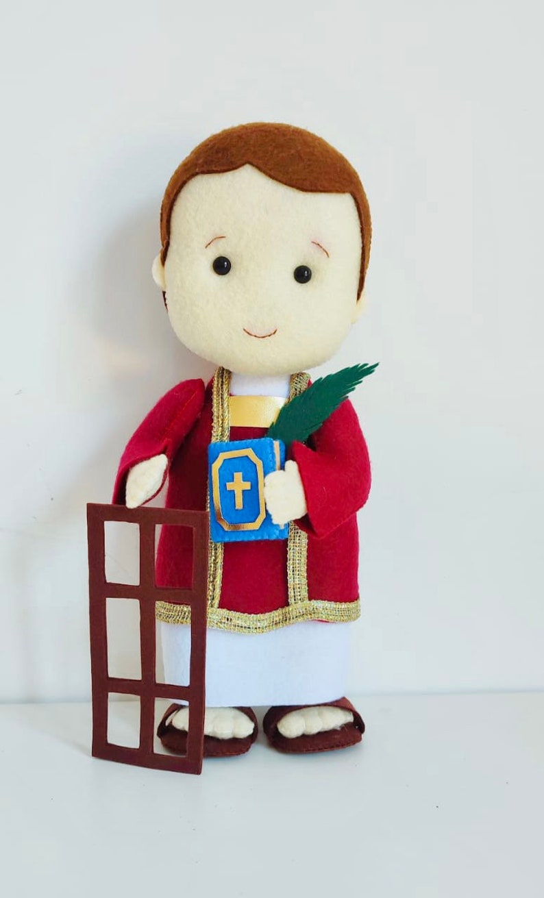 May include: A felt doll of a religious figure wearing a red robe with gold trim, holding a book with a cross on it and a palm branch. The doll is standing next to a brown felt cutout of a window.
