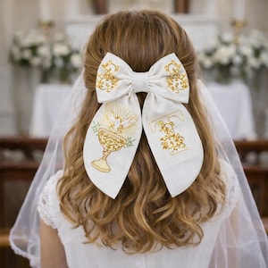 May include: A white hair bow with gold embroidery, depicting a chalice, dove, and the letter "T". The bow is worn in the hair, with a veil visible. The design is suitable for a religious ceremony.