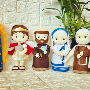 May include: A set of seven felt dolls representing various religious figures. The dolls are all dressed in traditional clothing and have smiling faces. The dolls are arranged in a row on a white brick background.