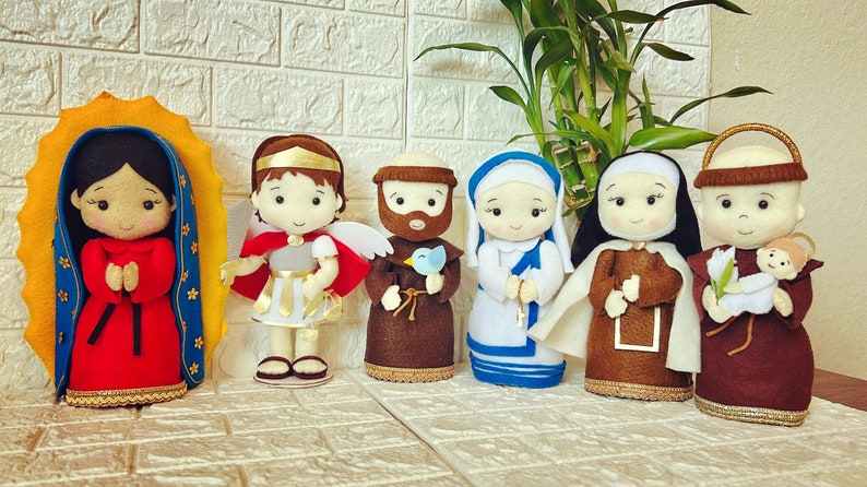 May include: A set of seven felt dolls representing various religious figures. The dolls are all dressed in traditional clothing and have smiling faces. The dolls are arranged in a row on a white brick background.