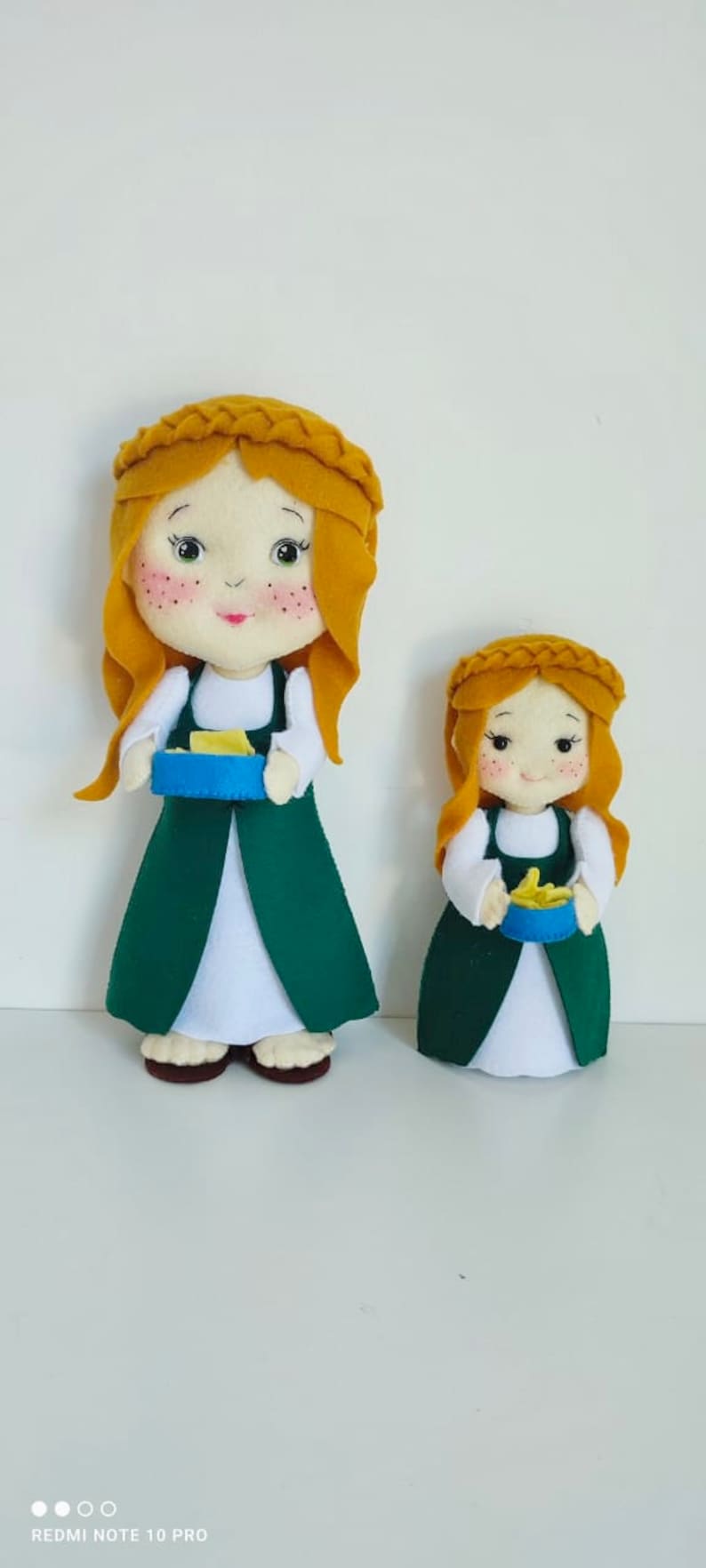 May include: Two felt dolls dressed in green dresses with white aprons and yellow hair. The dolls are holding blue bowls with yellow food inside.