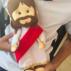 May include: A plush toy of Jesus wearing a white robe with a red sash and gold trim. The toy has brown hair, a brown beard, and a smiling face.