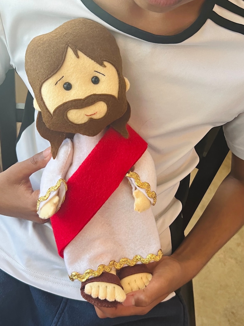 May include: A plush toy of Jesus wearing a white robe with a red sash and gold trim. The toy has brown hair, a brown beard, and a smiling face.