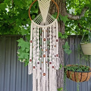 May include: A white macrame dream catcher with wooden beads hanging from a wooden hoop. The dream catcher is hanging from a tree branch.
