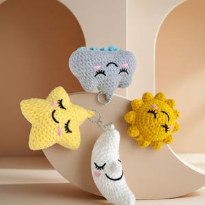 May include: A collection of crocheted celestial-themed ornaments. Includes a yellow star, a white crescent moon, a yellow sun, and a gray elephant, all with smiling faces and eyelashes. Each ornament is connected by a silver chain.