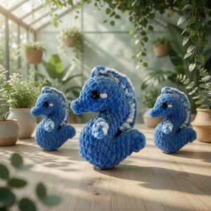 May include: Three handmade blue crochet seahorse figurines with white accents and black eyes are displayed on a wooden surface. The seahorses are in a well-lit, plant-filled environment, suggesting a nature-inspired theme.