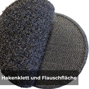 May include: Close-up of two dark grey circular hook-and-loop fastener pads. One pad has a fuzzy, textured surface, while the other has a more tightly woven, looped surface. The text "Hakenklett und Flauschfl&auml;che" is visible.