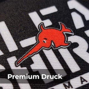 May include: Close-up of a black and white fabric featuring a red cartoon fish emblem. The text "Premium Druck" is at the bottom of the image. The fabric has a textured appearance.