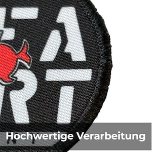 May include: A circular black patch with white lettering and a red graphic. The text includes the word "HEAT" and a number "1". The patch has a stitched border and the text "Hochwertige Verarbeitung" is visible at the bottom.