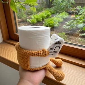 May include: A roll of white toilet paper is encased in a crocheted orange cover with arms and legs. A small, white crocheted piece with black lines is attached to the roll. The item is held in a hand.
