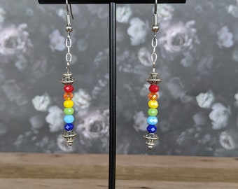 May include: Rainbow coloured glass bead earrings with silver accents hanging from a black earring stand.