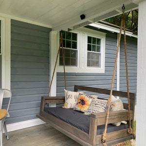May include: A wooden porch swing with a black cushion and pillows with a sunflower and text that reads "Be Kind, Be Grateful, Be Happy". The swing is hanging from the ceiling of a porch with a gray wooden siding.