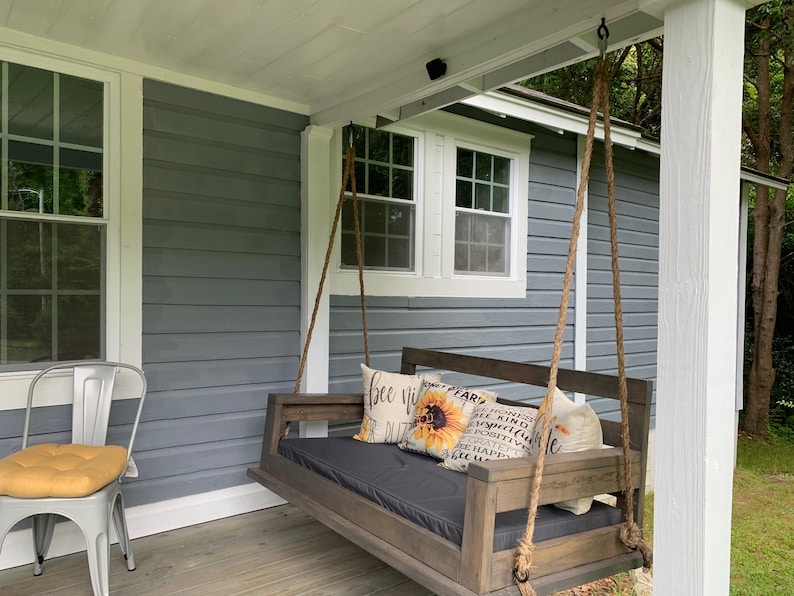 May include: A wooden porch swing with a black cushion and pillows with a sunflower and text that reads "Be Kind, Be Grateful, Be Happy". The swing is hanging from the ceiling of a porch with a gray wooden siding.