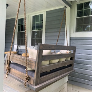 May include: A wooden swing bench with a gray cushion and pillows hanging from a porch ceiling. The swing is made of weathered wood with a dark stain. The porch is painted white with gray siding.