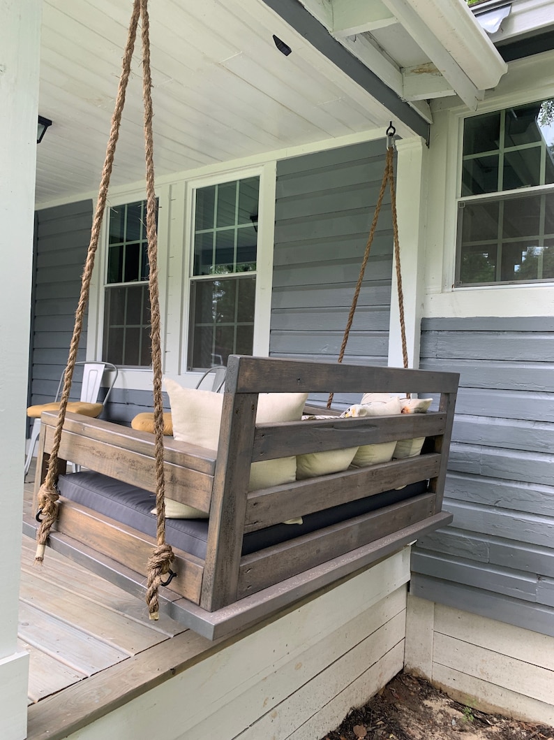 May include: A wooden swing bench with a gray cushion and pillows hanging from a porch ceiling. The swing is made of weathered wood with a dark stain. The porch is painted white with gray siding.