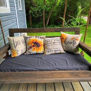 May include: A wooden porch swing with a black cushion and four throw pillows. The pillows have a variety of designs, including a bee and sunflower, and text that says "bee kind" and "home where your heart is".