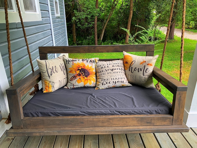 May include: A wooden porch swing with a black cushion and four throw pillows. The pillows have a variety of designs, including a bee and sunflower, and text that says "bee kind" and "home where your heart is".