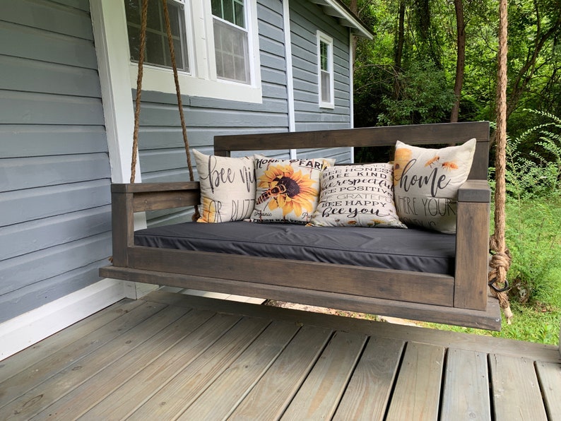 May include: A wooden porch swing with a black cushion and three throw pillows. The pillows have a sunflower and bee design and the text "bee happy" and "home".