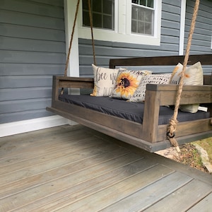 May include: A wooden porch swing with a black cushion and pillows with sunflower and floral patterns. The swing is hanging from a porch with gray siding and a wooden floor.