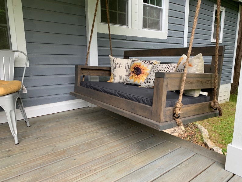 May include: A wooden porch swing with a black cushion and pillows with sunflower and floral patterns. The swing is hanging from a porch with gray siding and a wooden floor.